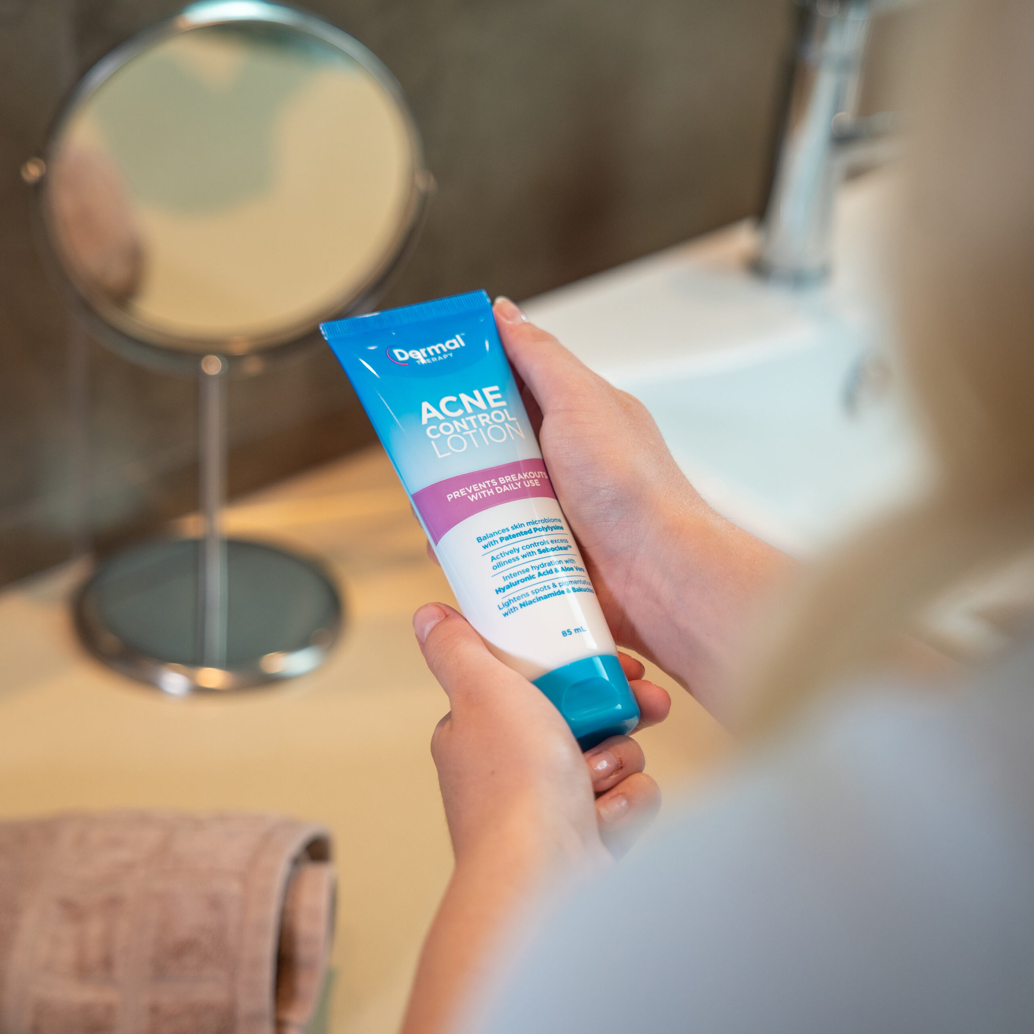 A woman holding a Dermal Therapy Acne Control Lotion tube in the bathroom, with a shaving mirror in the background.