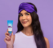 Rojin Torabi holding a Dermal Therapy Overnight Lip Repair Mask, with a sleep mask on her forehead on a purple background. 