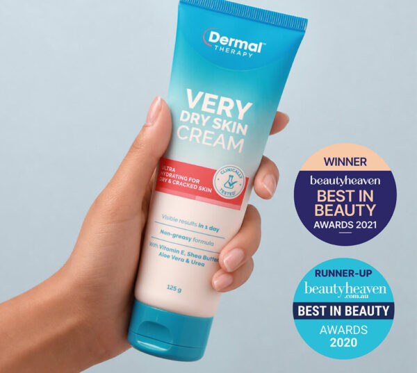 Hand holding a tube of Dermal Therapy Very Dry Skin Cream against a light blue background with award badges displayed beside the product