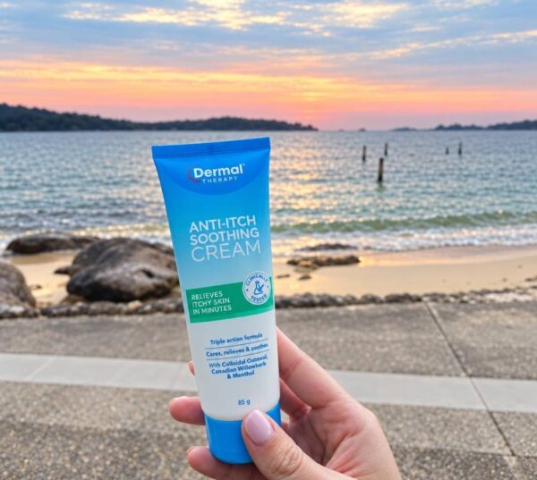 Hand holding a tube of Dermal Therapy Anti-Itch Soothing Cream in front of a beach and ocean backdrop at sunset.